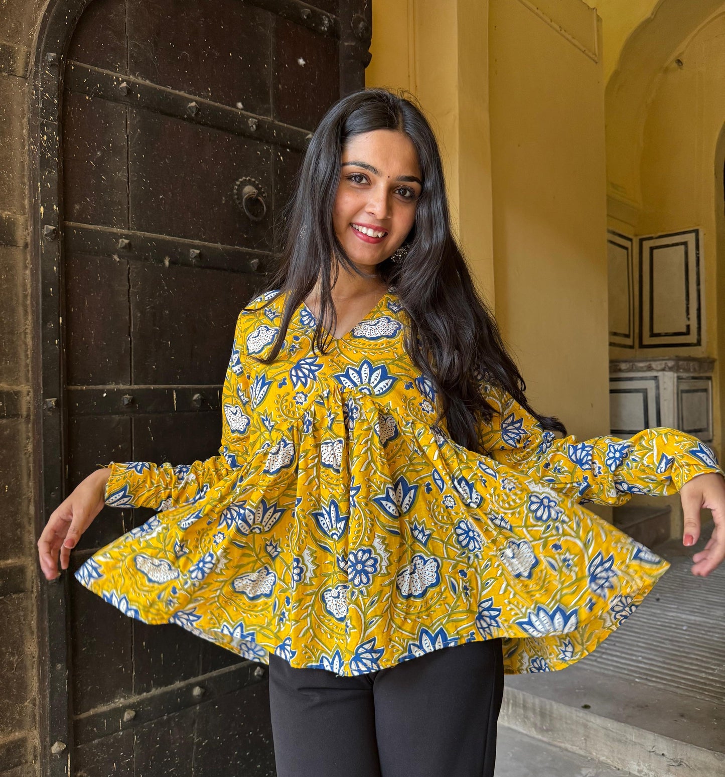 A woman standing in an archway, wearing a yellow kurti with floral designs and full sleeves.