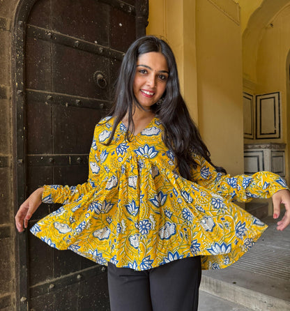 A woman standing in an archway, wearing a yellow kurti with floral designs and full sleeves.