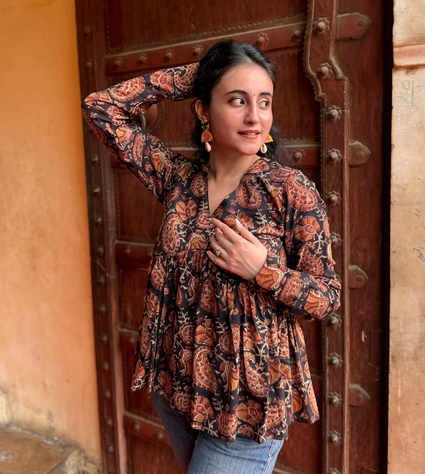 A woman standing with one hand against a door, wearing a black and brown floral kurti with 3/4 length sleeves and a V-neck.