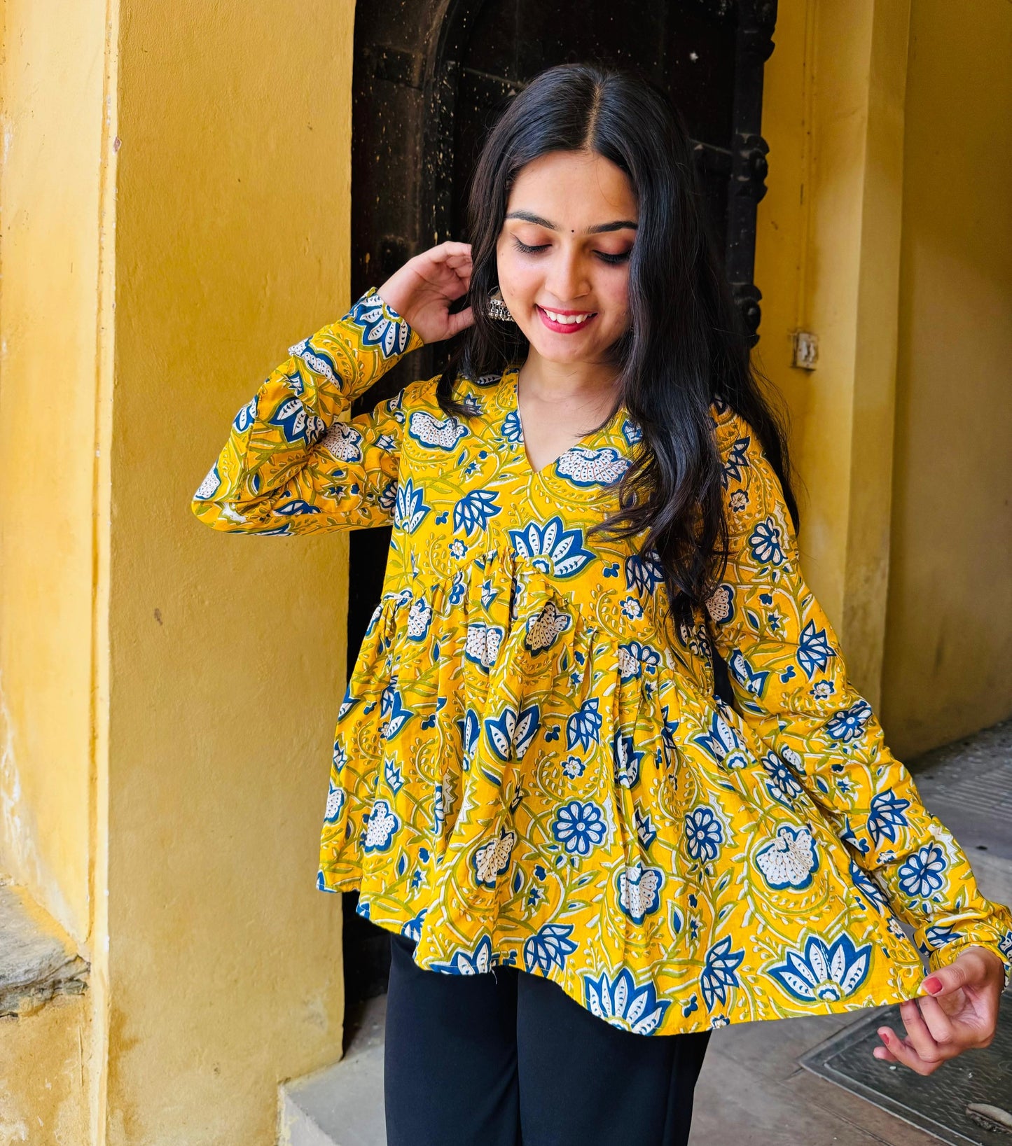 A woman standing in a sunny doorway wearing a yellow kurti with floral patterns and full sleeves.