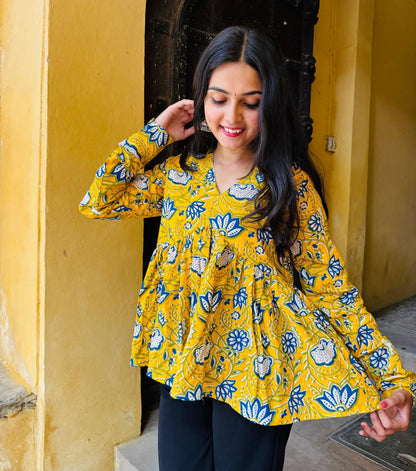 A woman standing in a sunny doorway wearing a yellow kurti with floral patterns and full sleeves.