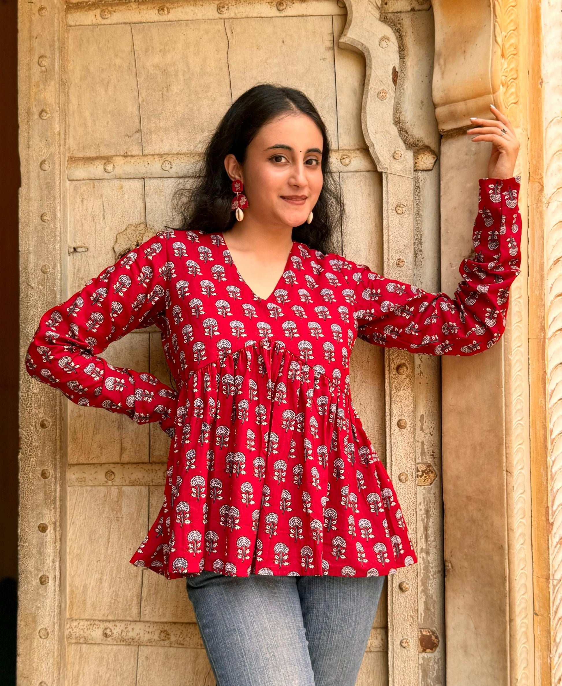 A woman wearing a red kurti with a floral pattern, standing in front of a wooden door.