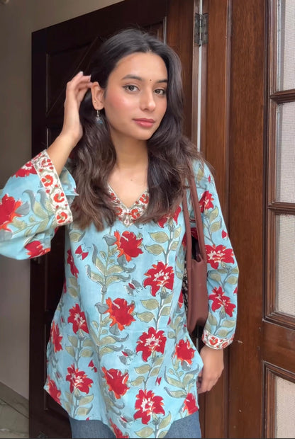 Woman wearing a floral blouse with red and green flowers in front of a wooden door.