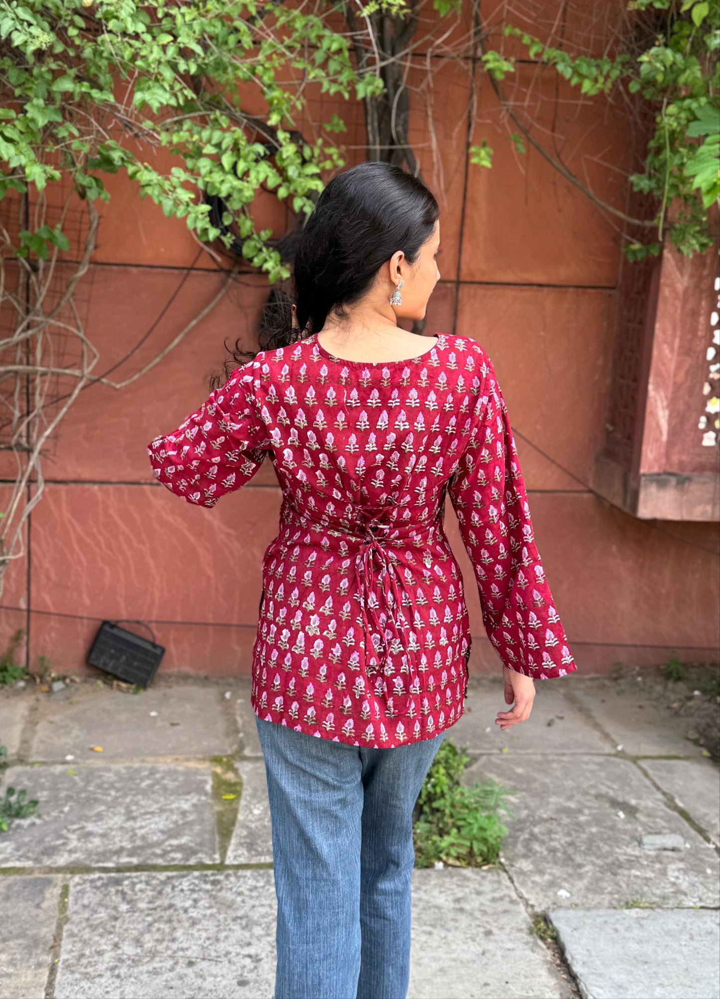 Person wearing a red patterned shirt and blue jeans standing in front of a red wall with greenery.
