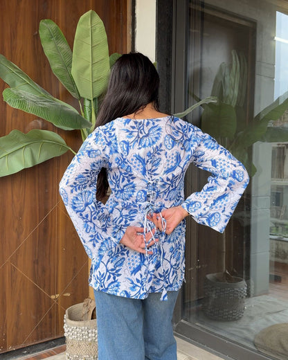 Woman wearing a blue floral blouse standing in front of a wooden door with plants.