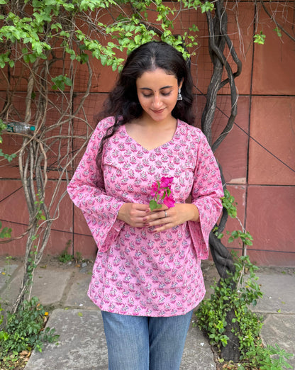 Woman wearing a pink floral blouse holding flowers against a red wall and green plants.