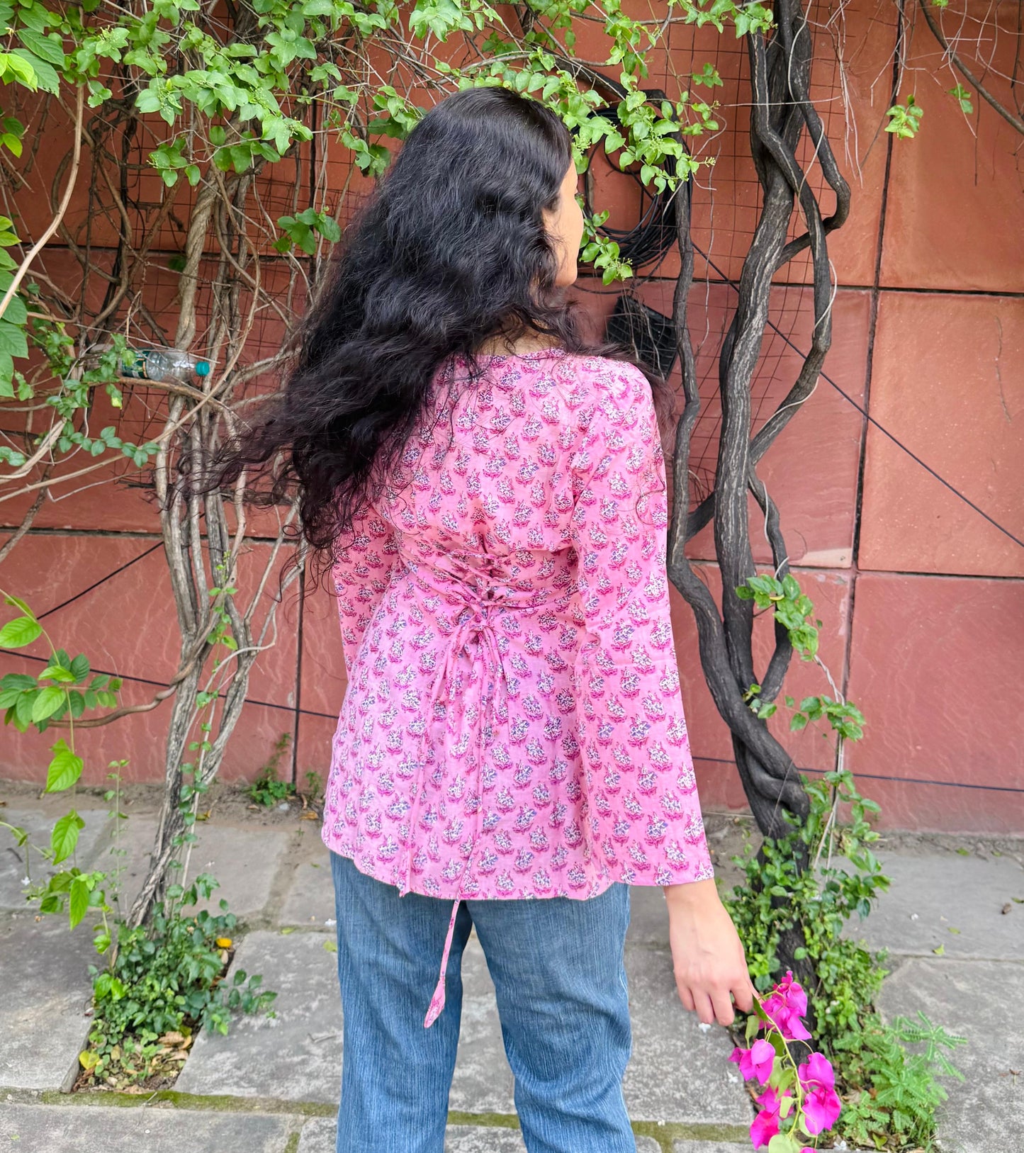 Person wearing a pink floral jacket and blue jeans standing near a red wall with greenery.