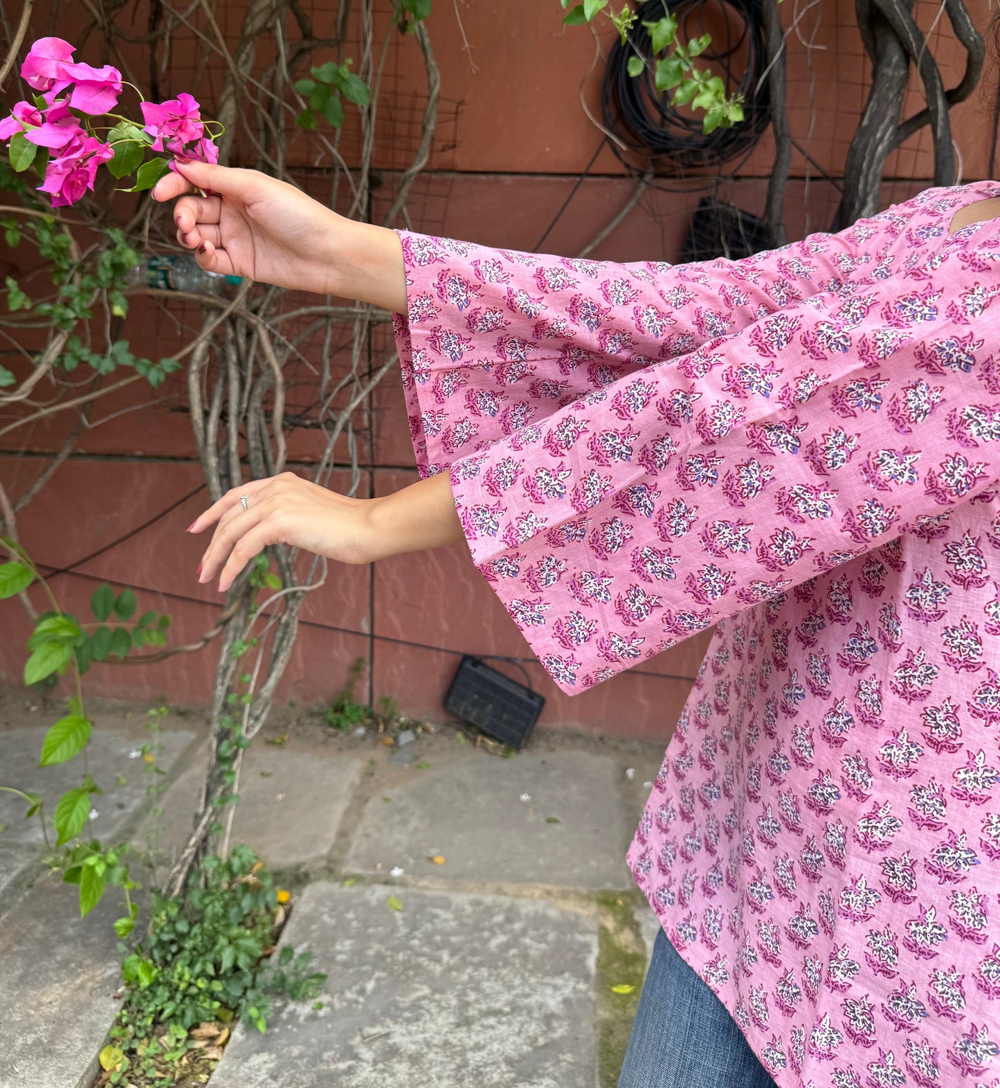 Person wearing a pink floral dress holding a pink flower in an outdoor setting.