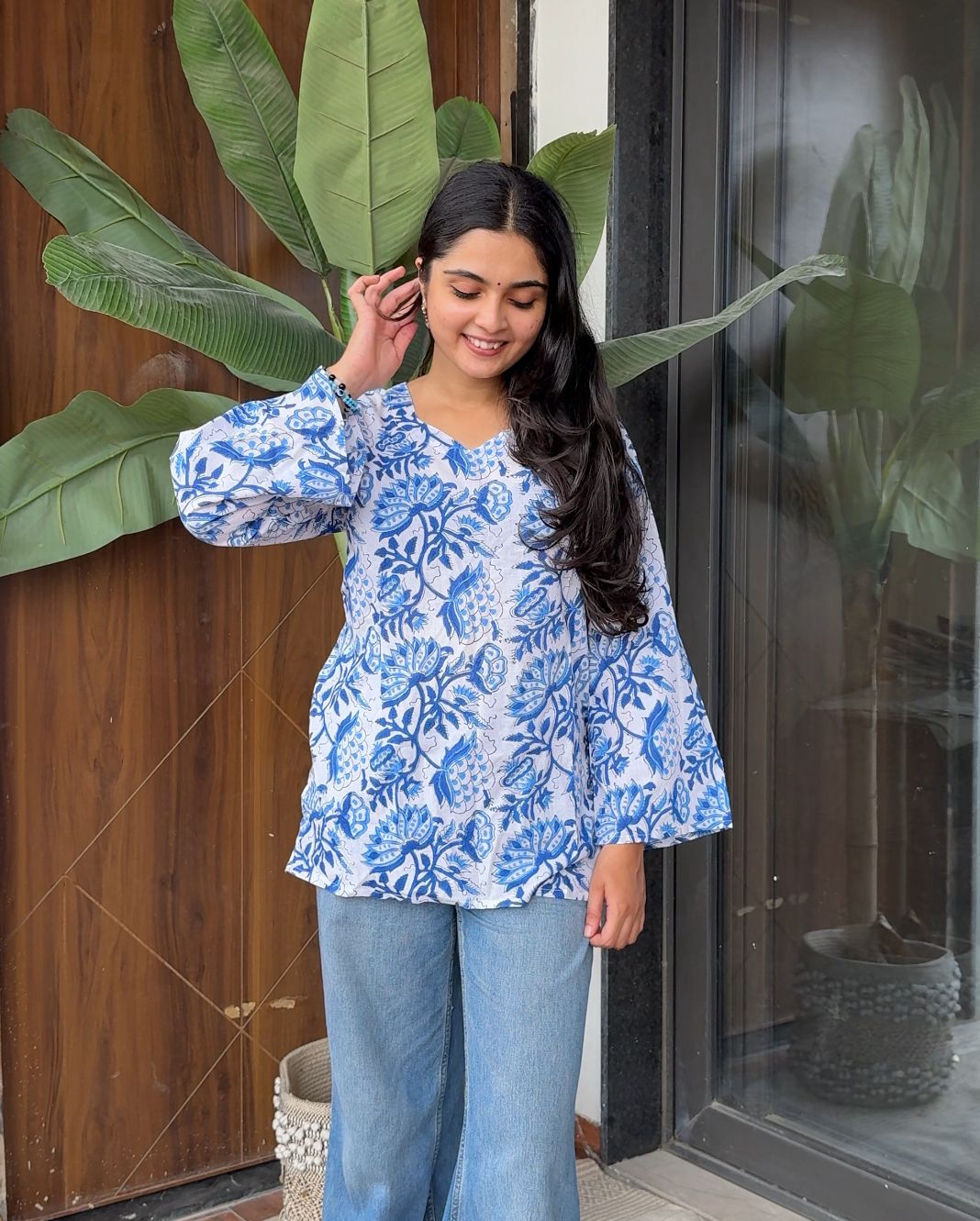 Woman wearing a blue floral top and jeans standing indoors with plants in the background