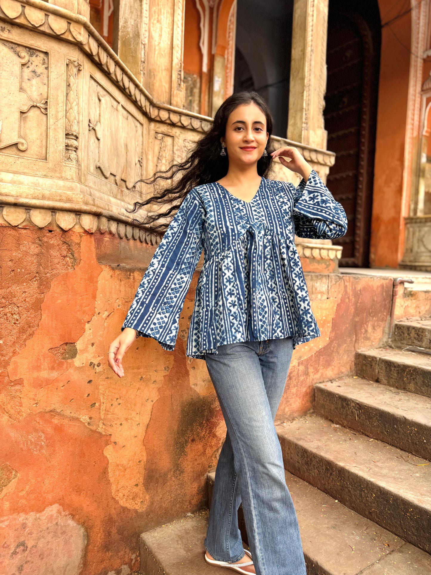 Woman in a blue patterned top and jeans standing on steps with an ornate wall in the background