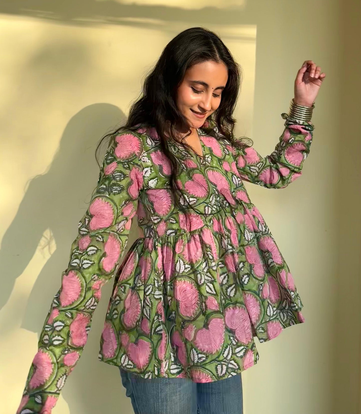 A woman wearing a floral hand-block printed cotton top in pink and green hues, with long sleeves and a round neckline.