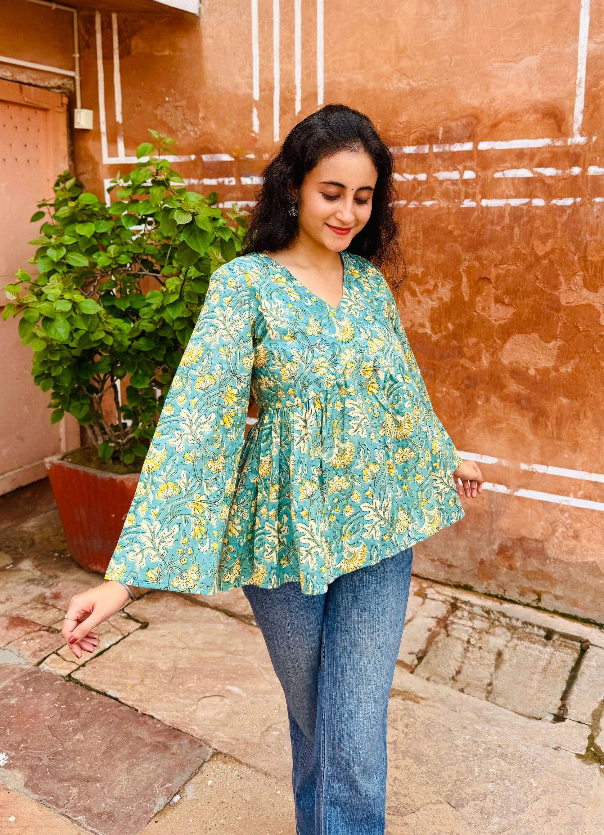 Woman wearing a teal floral blouse and blue jeans standing against a textured wall.