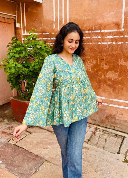 Woman wearing a teal floral blouse and blue jeans standing against a textured wall.