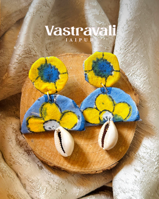Pair of colorful earrings with cowrie shells on a wooden board, branded 'Vastravali'.