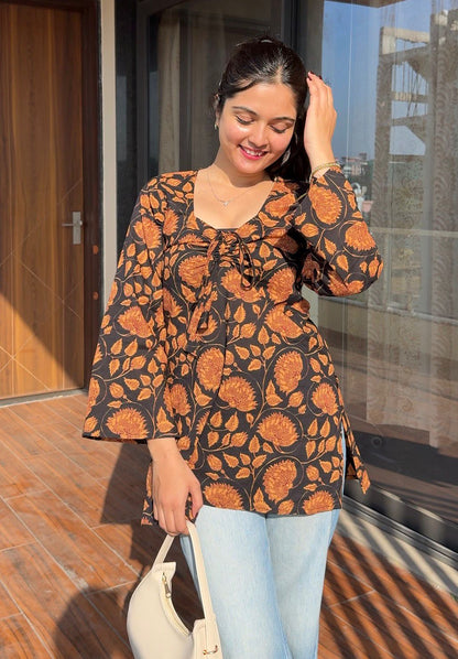 Woman wearing a patterned top and light blue jeans, standing on a wooden floor with a glass door in the background.