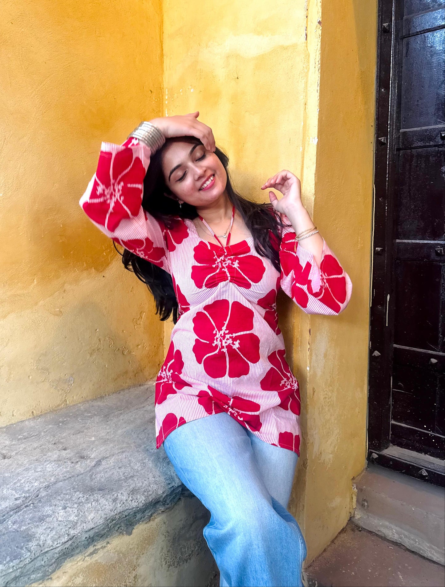 Woman in a red floral top and blue pants leaning against a yellow wall.