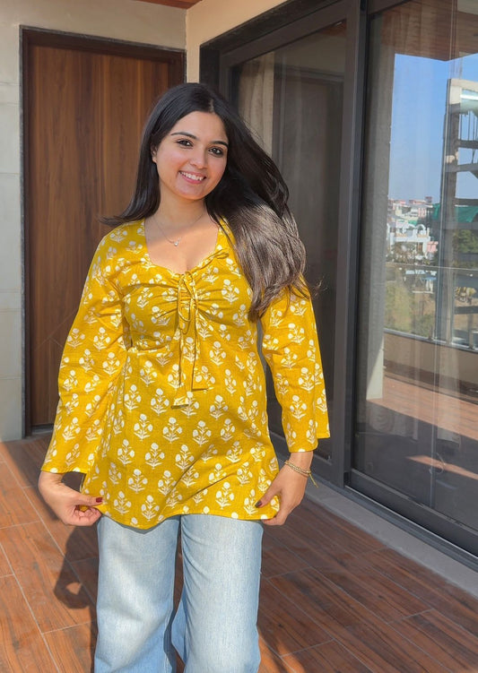 Woman wearing a yellow floral blouse and light blue jeans standing indoors.
