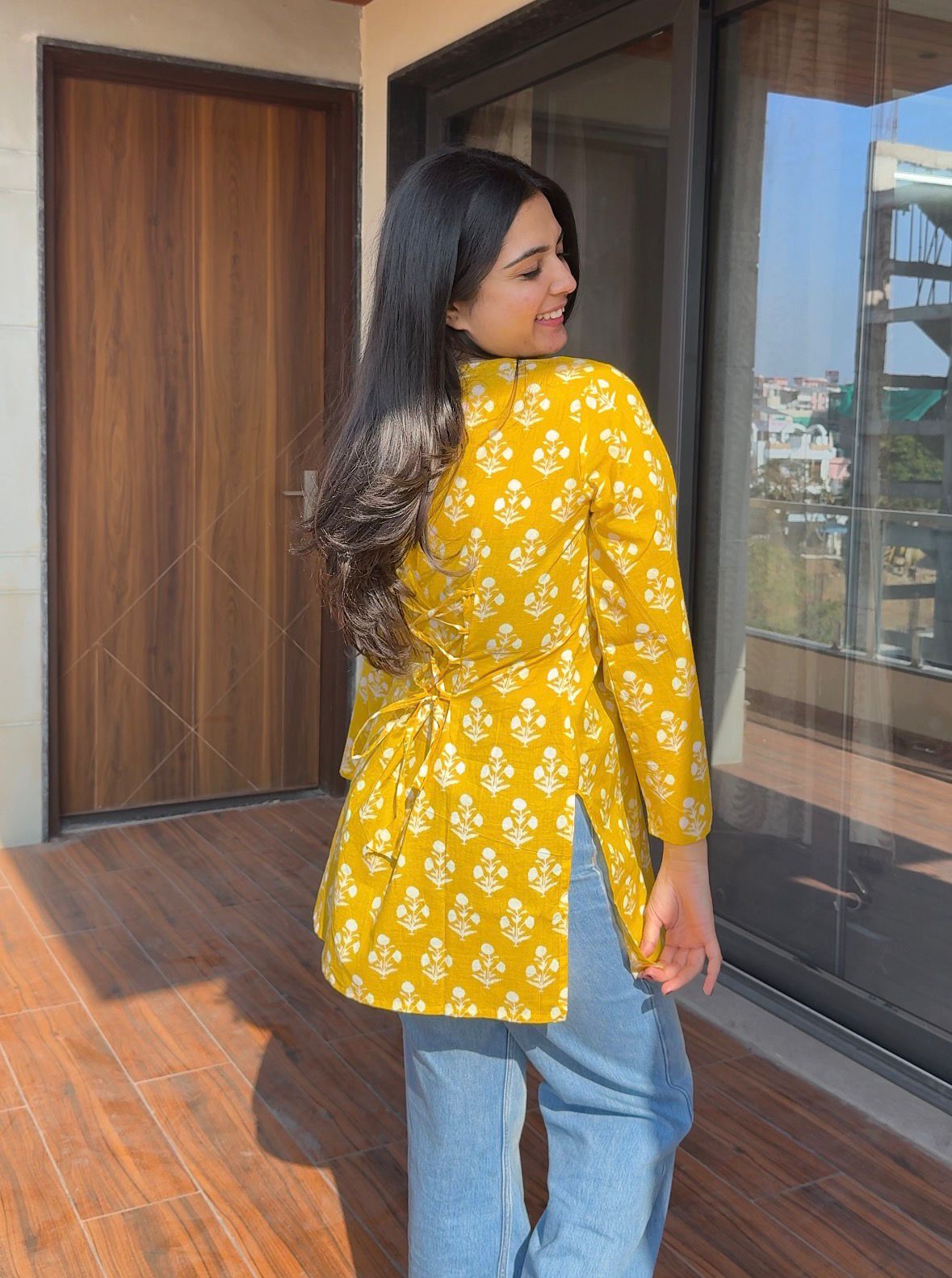 Woman wearing a yellow floral blouse and blue jeans standing on a wooden floor.