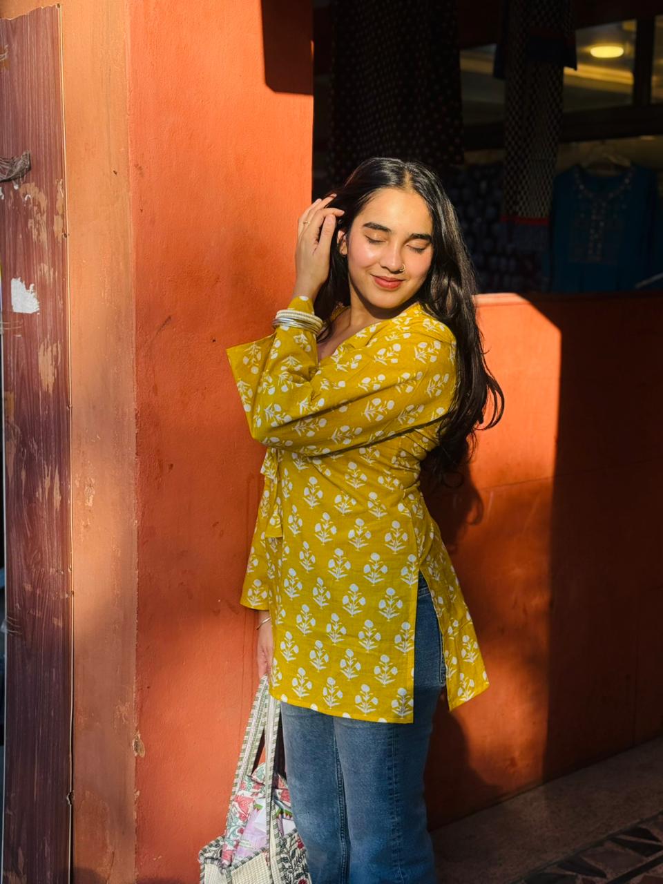 Woman in a yellow floral blouse and blue jeans standing against an orange wall.