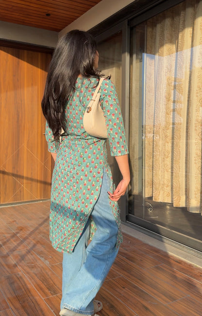 Woman in a green patterned shirt and blue jeans walking on a wooden floor.