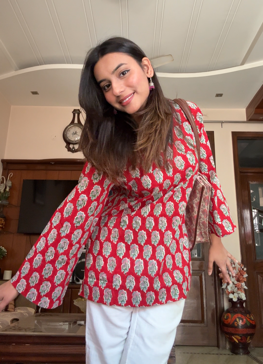 A woman standing in an indoor setting wearing a red and white hand-block printed short kurti with flared sleeves and white pants.