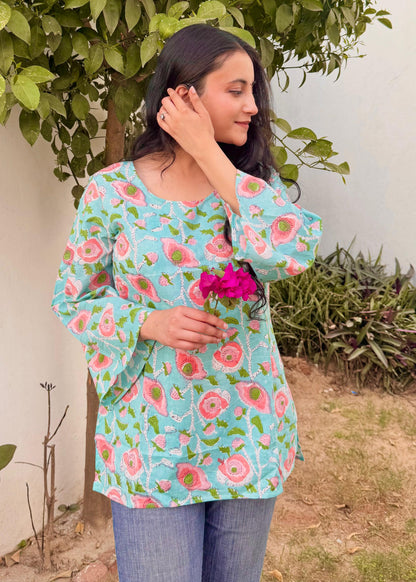 A woman standing outdoors wearing a blue short kurti with hand-block print designs, holding a pink flower.