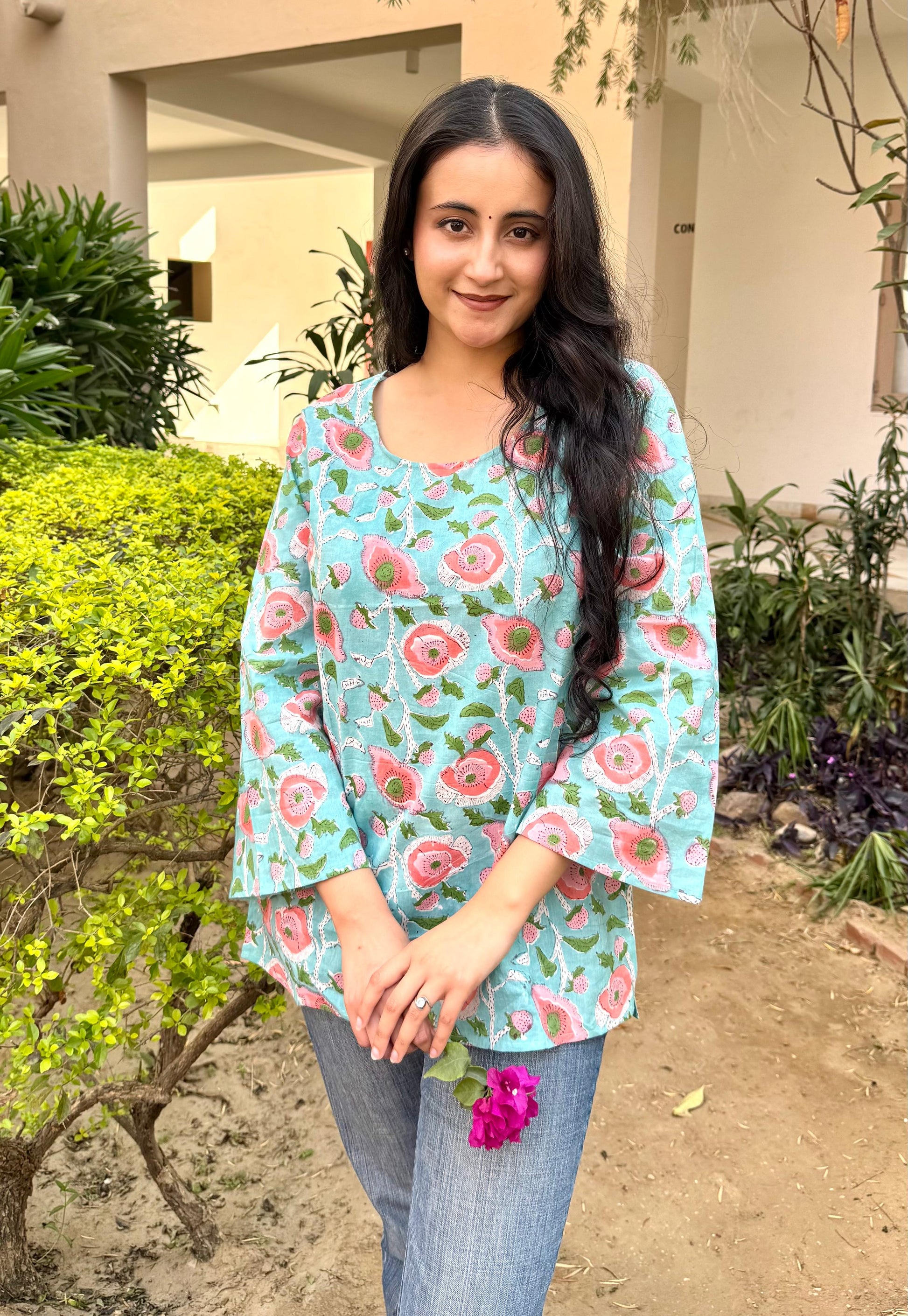 A woman standing outdoors in a garden wearing a blue short kurti with hand-block print designs.
