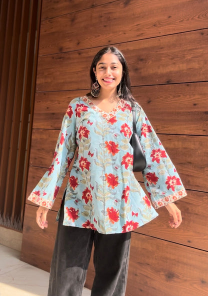 A woman standing against a wooden background, wearing a floral hand-block printed kurti with flared sleeves.