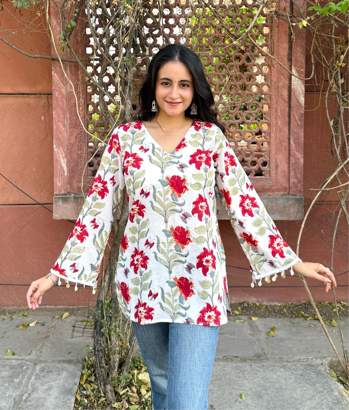 A woman standing outdoors wearing a white short kurti with red hand-block prints and flared sleeves.