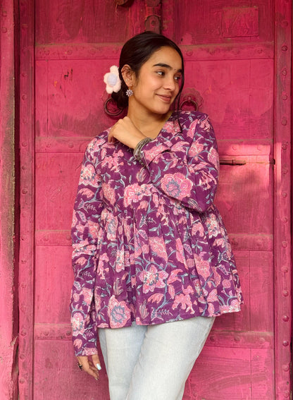 A woman standing in front of a pink door, wearing a purple floral kurti with flared sleeves and a round neckline, paired with white leggings.