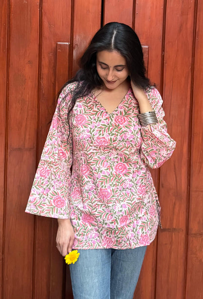 A woman wearing a pink floral short kurti with flared sleeves, standing in front of a wooden background.
