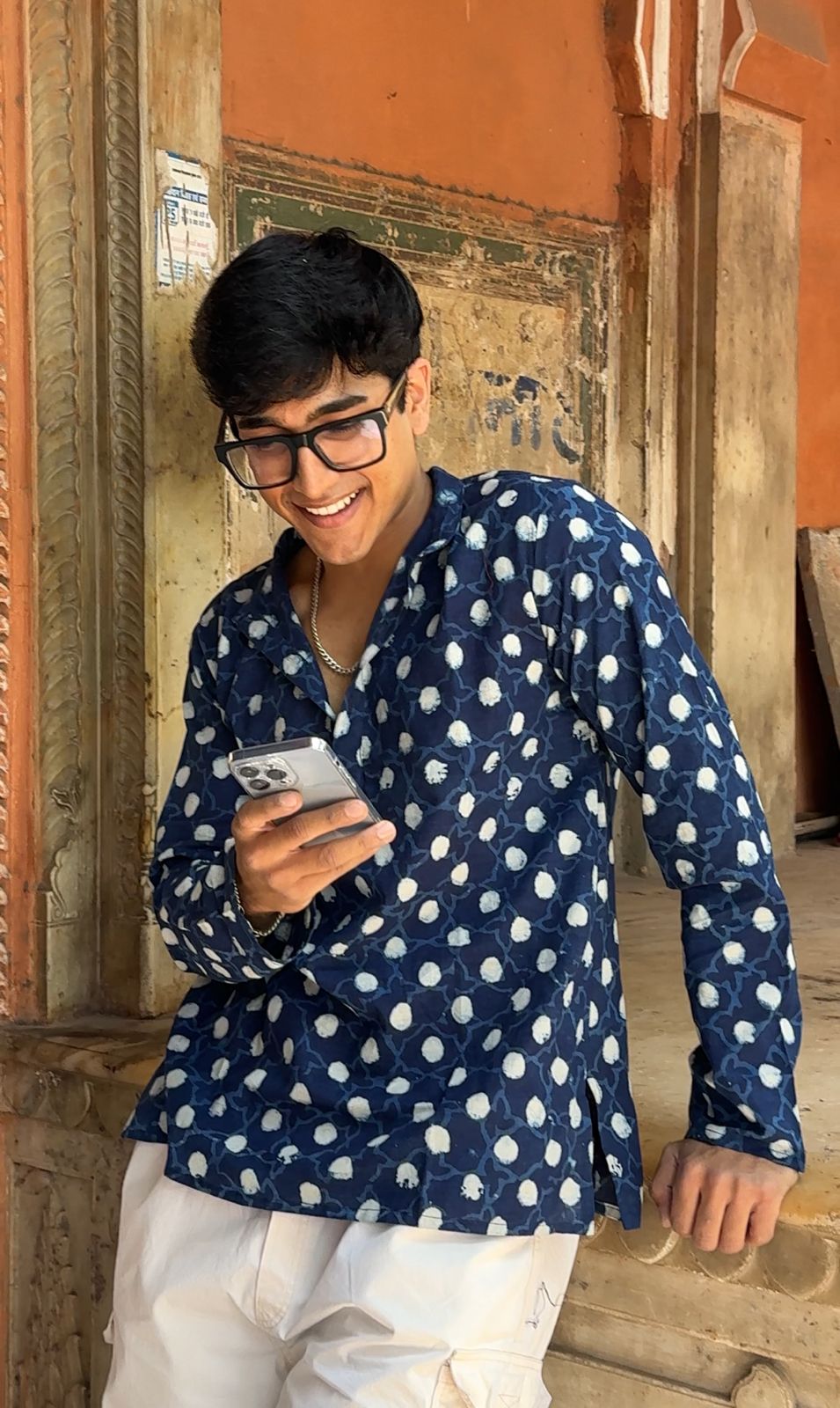 Man wearing a blue polka dot shirt and glasses, holding a phone, standing against an orange wall.