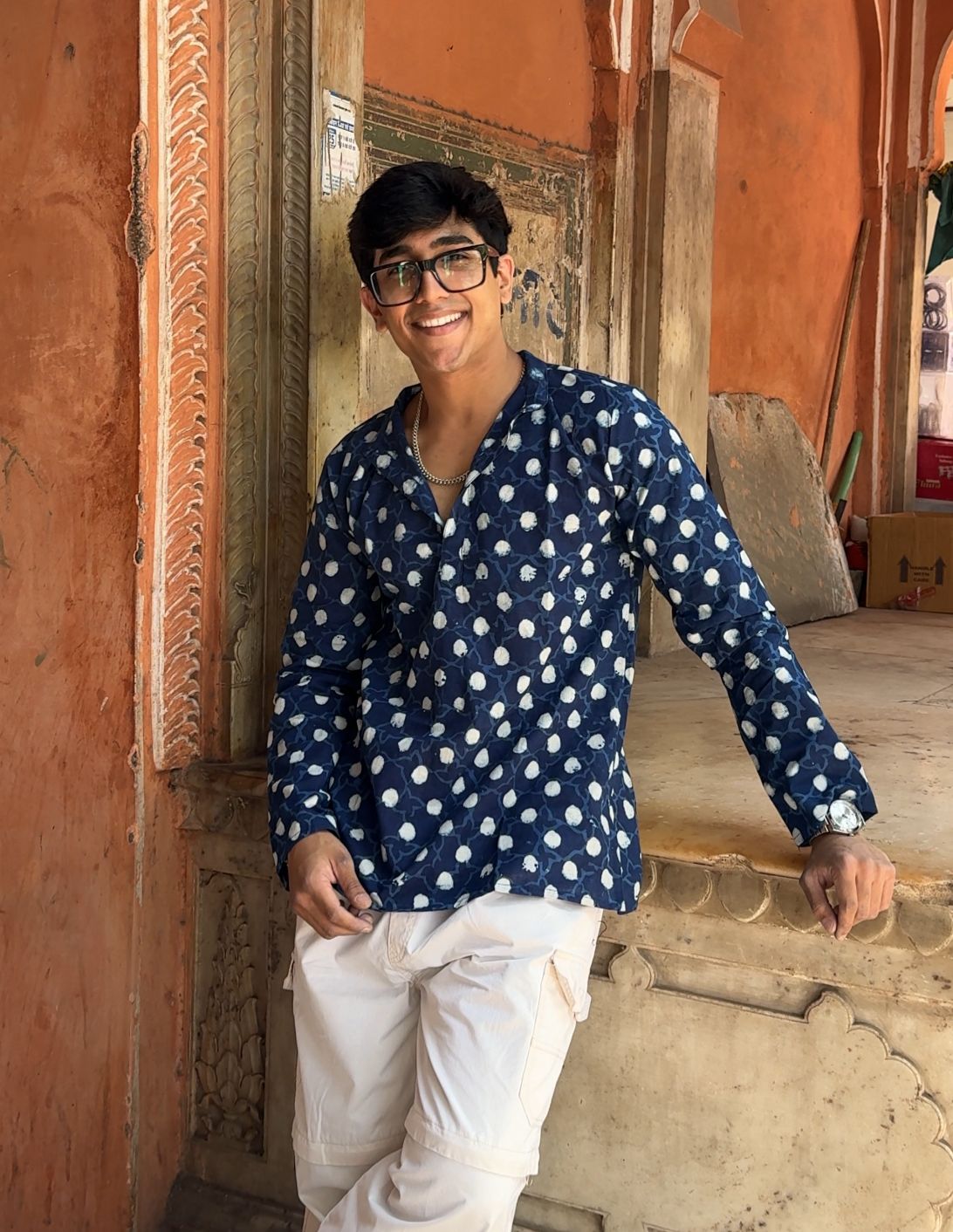 Man wearing a blue polka dot shirt and white pants standing in an ornate doorway.