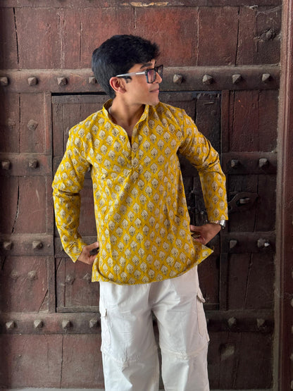 Person wearing a yellow patterned shirt and white pants standing against a wooden door.