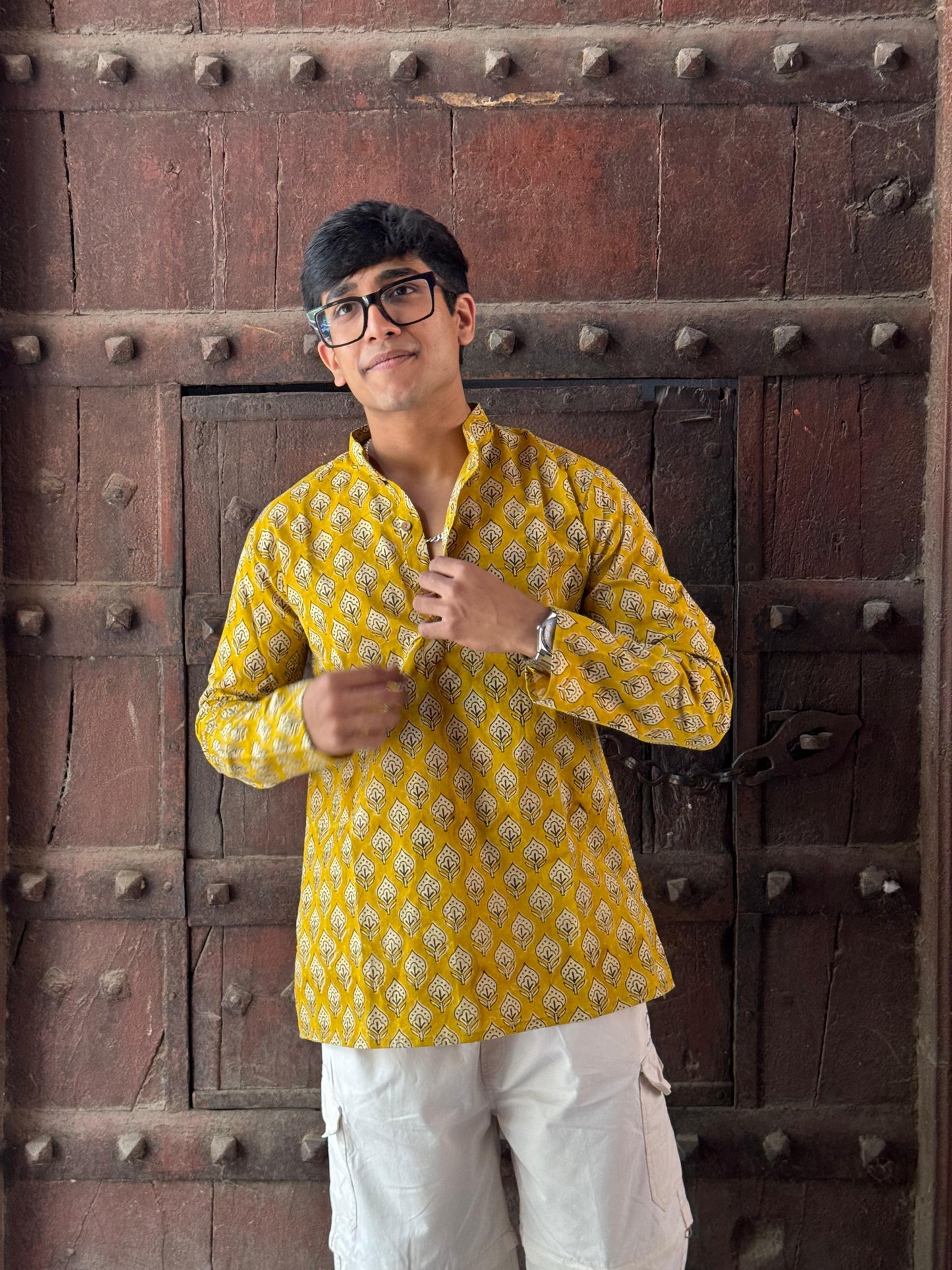 Man wearing a yellow patterned shirt and white pants standing in front of a textured wall.
