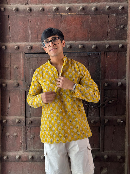 Man wearing a yellow patterned shirt and white pants standing in front of a textured wall.