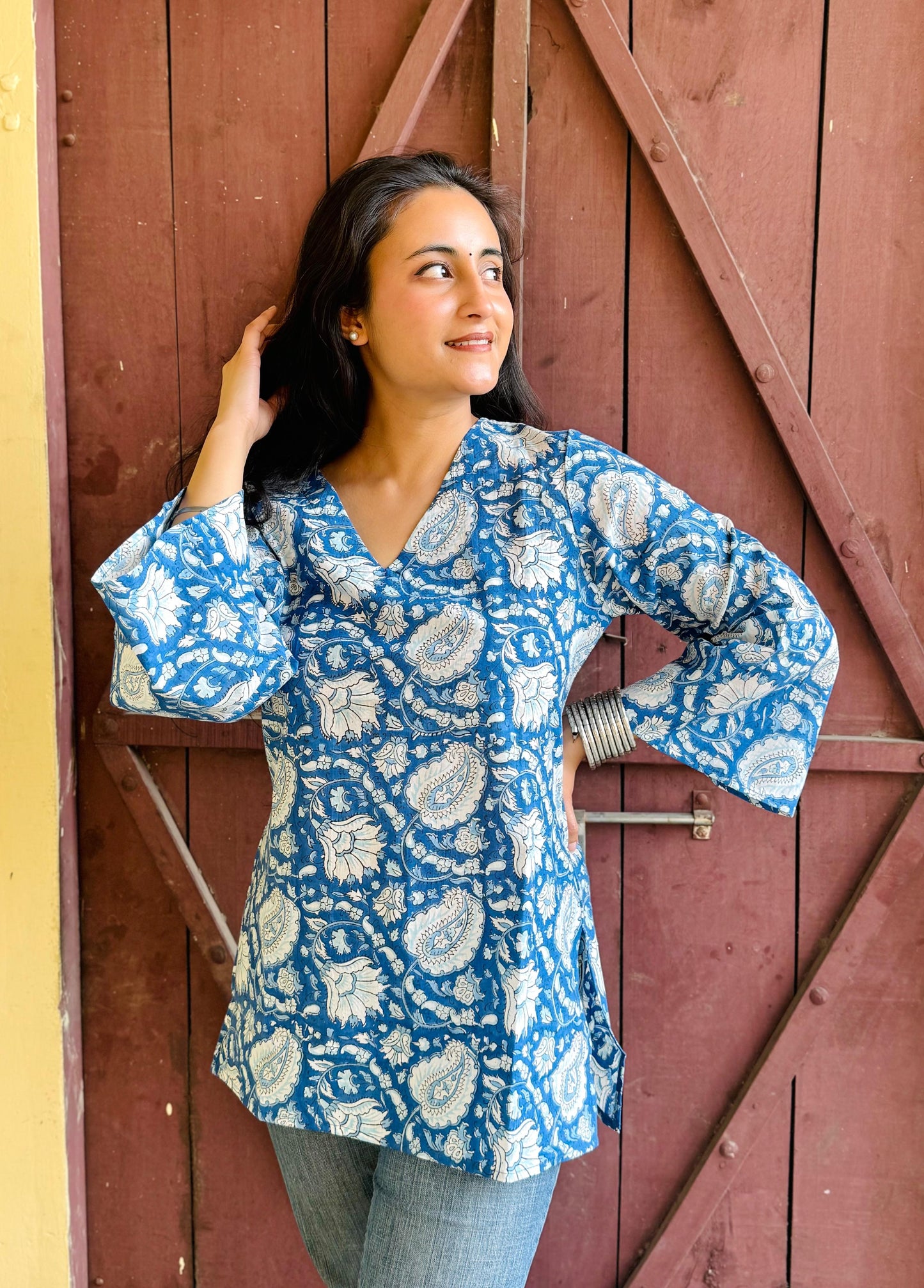 A woman wearing a blue and white hand-block printed short kurti with flared sleeves, standing against a wooden backdrop.