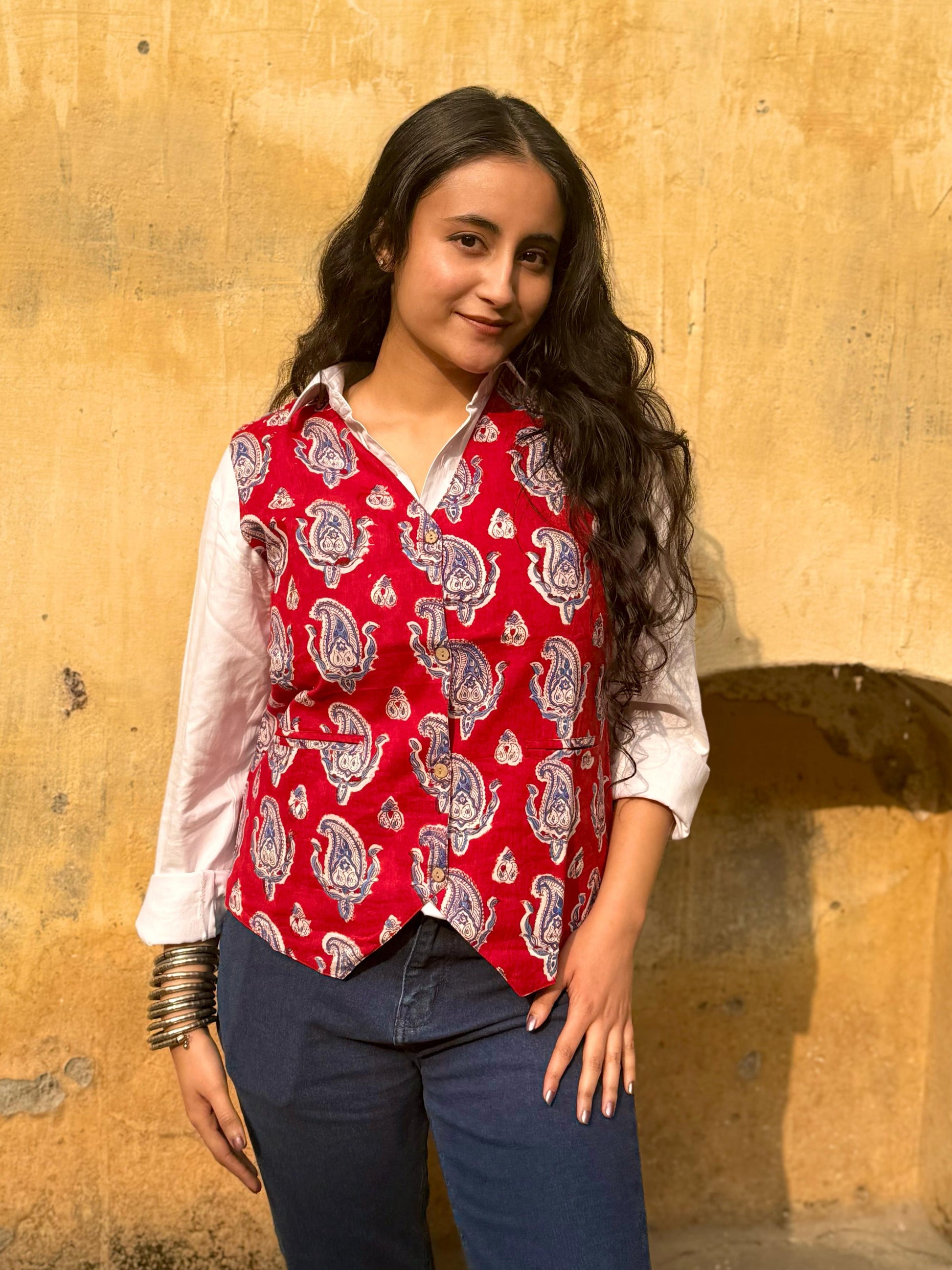A woman standing against a yellow wall, wearing a red and white floral patterned vest top with 3/4 sleeves, paired with blue jeans.