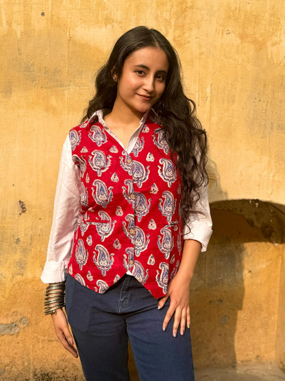 A woman standing against a yellow wall, wearing a red and white floral patterned vest top with 3/4 sleeves, paired with blue jeans.
