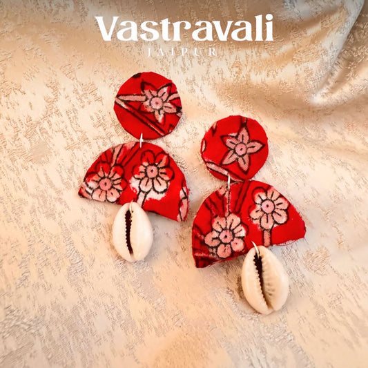 A pair of hand-made fabric earrings with a red floral pattern and shell accents, displayed on a beige background.