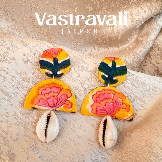 A pair of handmade fabric earrings with floral patterns and shell accents, displayed on a textured background.