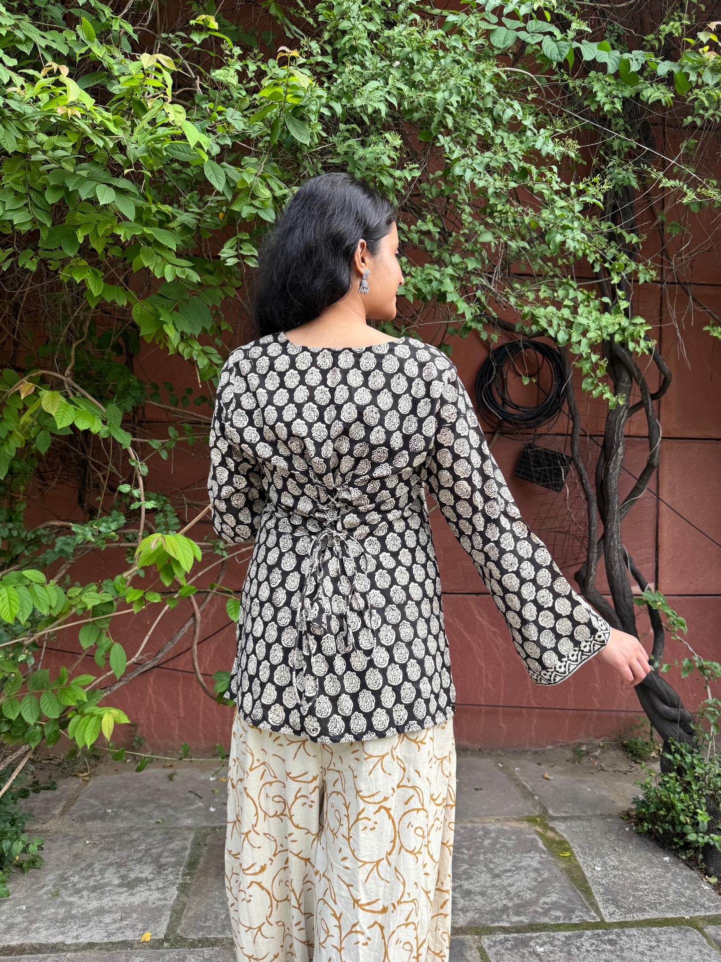 Woman wearing a black and white patterned top with long sleeves and light-colored pants, standing outdoors against a red wall with greenery.