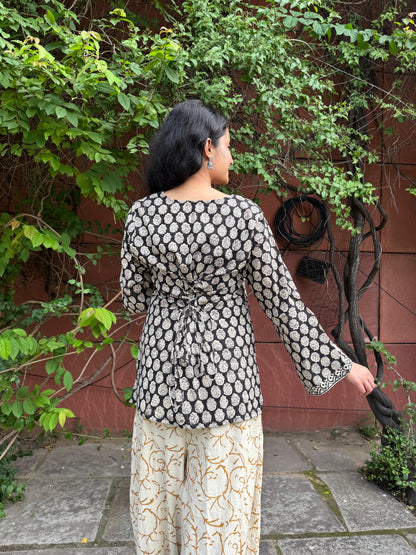 Woman wearing a black and white patterned top with long sleeves and light-colored pants, standing outdoors against a red wall with greenery.
